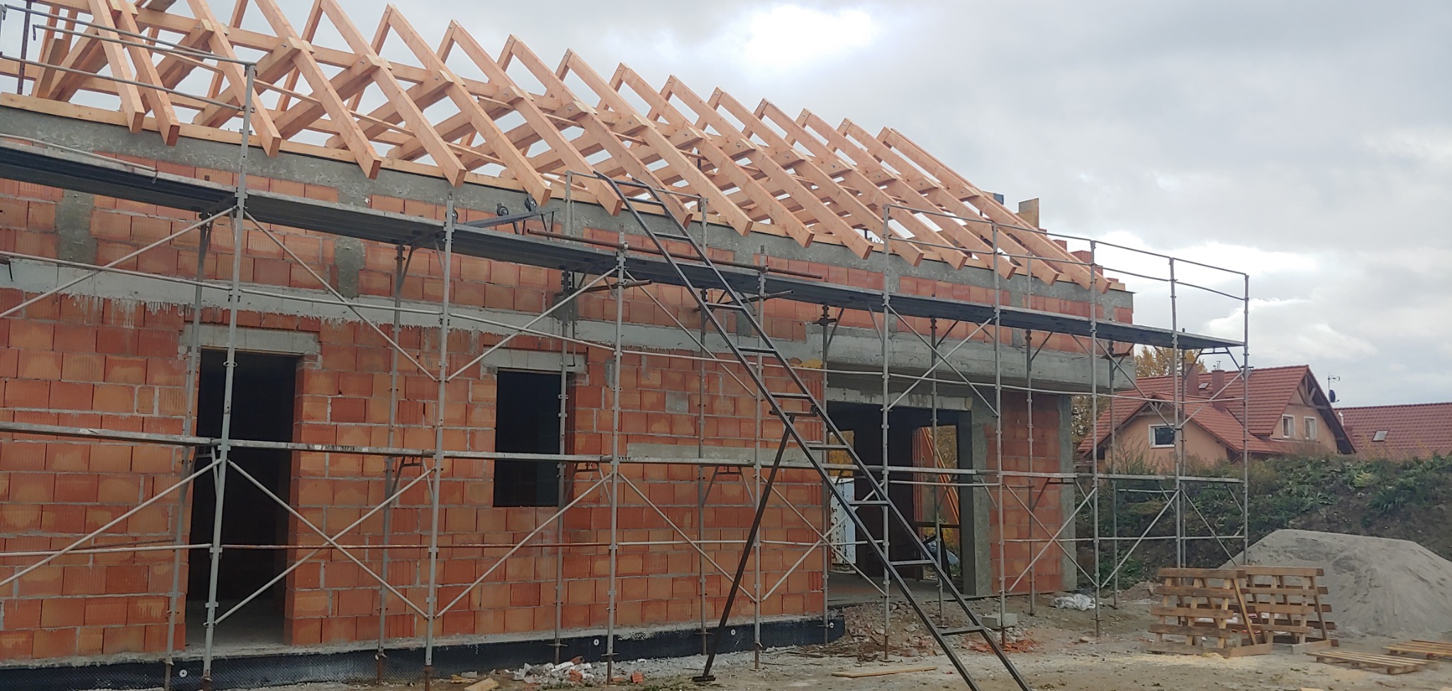 Budowa domu jednorodzinnego w trakcie wznoszenia więźby dachowej, widoczne rusztowania, ściany z czerwonej cegły i elementy drewniane konstrukcji dachu.