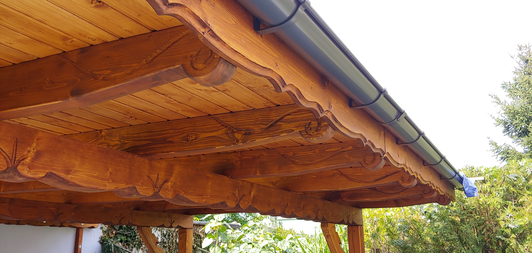Drewniana konstrukcja dachu z ozdobnymi wykończeniami i zamontowaną rynną w kolorze antracytowym, widok od dołu na tle zieleni.
