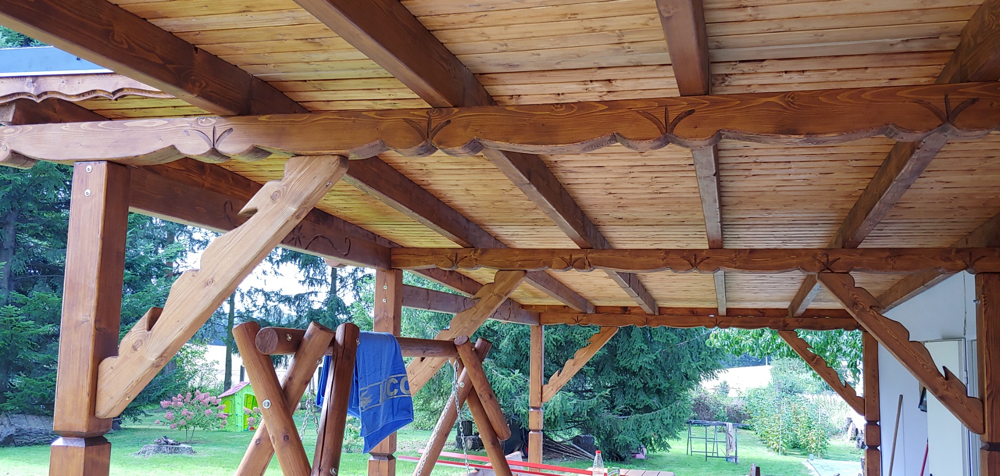Drewniana konstrukcja z dachem z desek, widoczne zdobienia krawędzi dachu i słupów, w tle zieleń ogrodu i huśtawka.