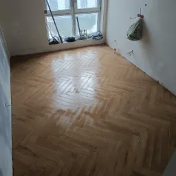 Ułożona w jodełkę, drewnopodobna podłoga z płytek, częściowo mokra, w trakcie prac wykończeniowych w pomieszczeniu z oknem i widocznymi instalacjami elektrycznymi.