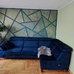 Salon z niebieską narożną kanapą, parkietem w jodełkę i ścianą z geometrycznym wzorem w odcieniach szarości i niebieskiego, widok na okno i rośliny doniczkowe.