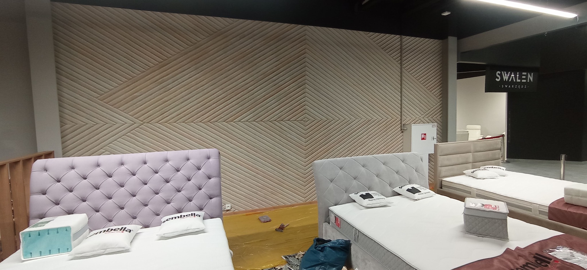 Wystawa łóżek z tapicerowanymi zagłówkami w salonie meblowym, na ścianie dekoracyjna okładzina z drewnianych listew, w tle widoczny szyld salonu 'SWALEN'.