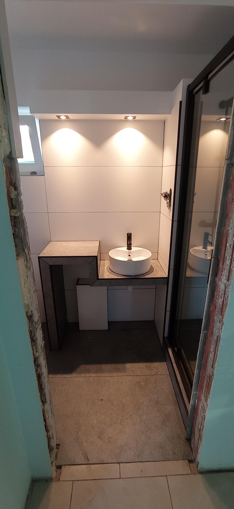 Nowoczesna łazienka z okrągłą umywalką nablatową i blatem z szarego kamienia, oświetlona punktowo, widok na kabinę prysznicową z czarnymi ramami, w trakcie prac wykończeniowych, z widocznymi...