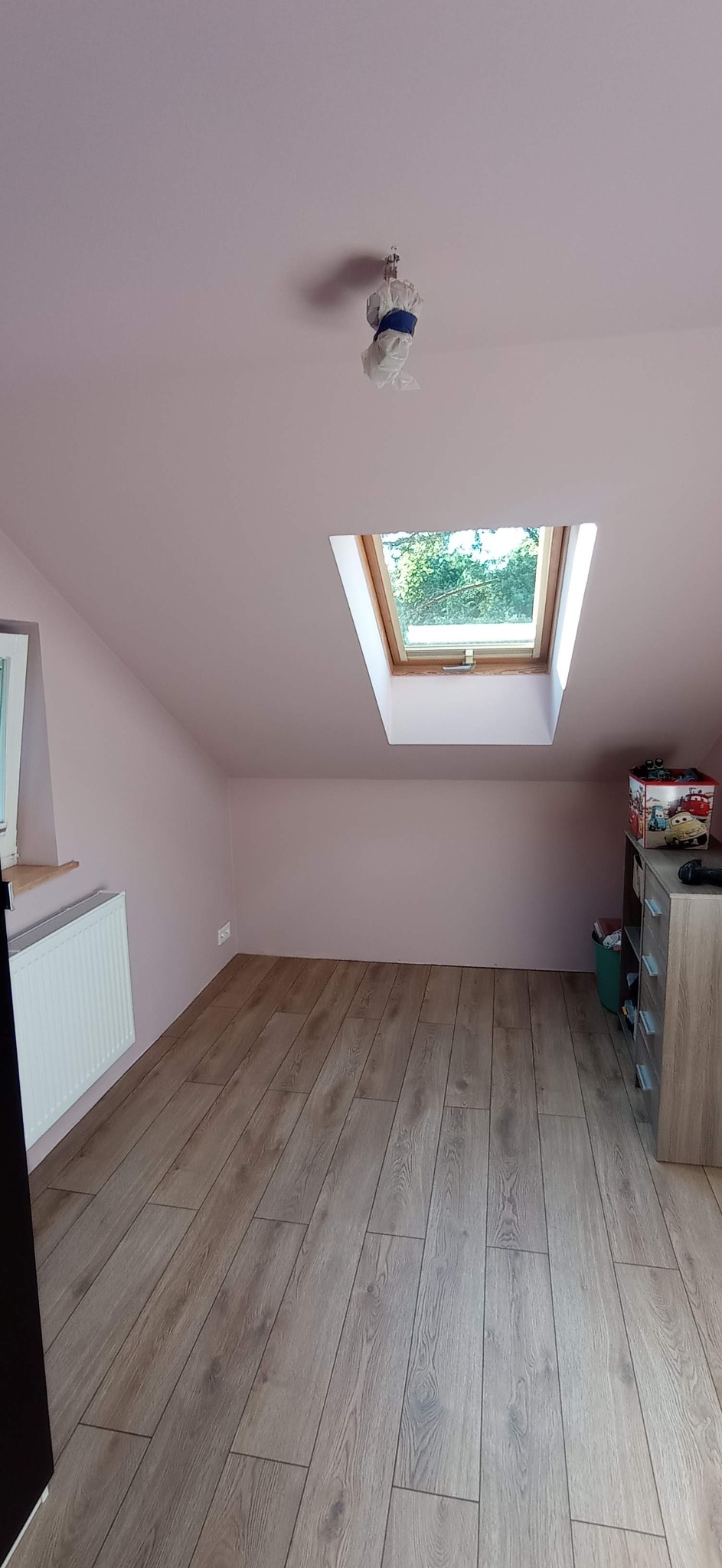 Poddasze pomalowane na różowo z drewnianą podłogą, okno dachowe z widokiem na zieleń, grzejnik pod oknem, zabezpieczona lampa sufitowa.