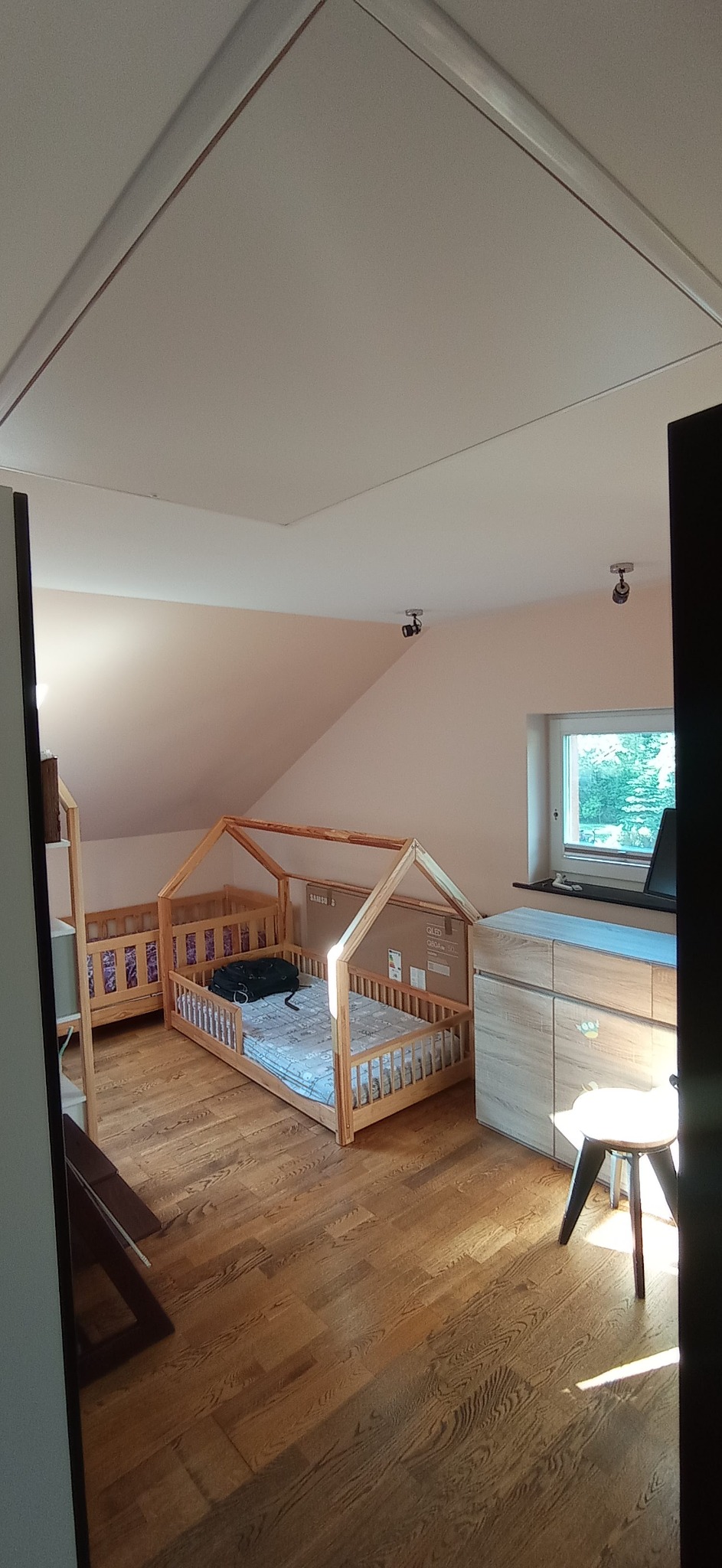 Pokój dziecięcy na poddaszu ze skośnym sufitem, drewnianą podłogą i dwoma łóżkami w kształcie domków, z których jedno jest częściowo zasłonięte kartonem po telewizorze.
