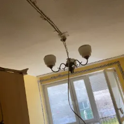 Pokój z żółtymi ścianami, widoczne listwy przypodłogowe z kablami, okno z otwartym skrzydłem, oraz wisząca lampa z trzema kloszami i odsłoniętym przewodem.