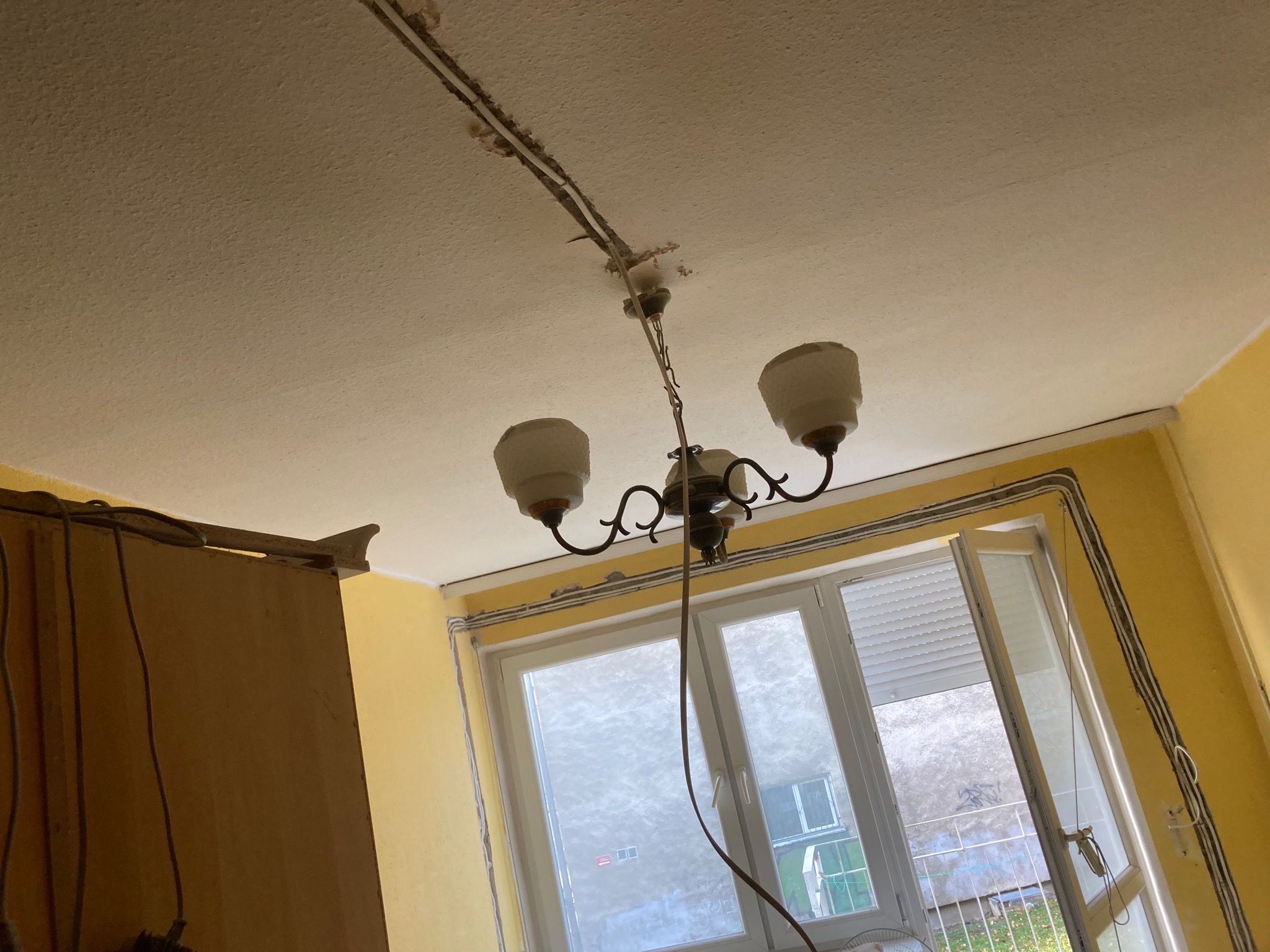 Pokój z żółtymi ścianami, widoczne listwy przypodłogowe z kablami, okno z otwartym skrzydłem, oraz wisząca lampa z trzema kloszami i odsłoniętym przewodem.