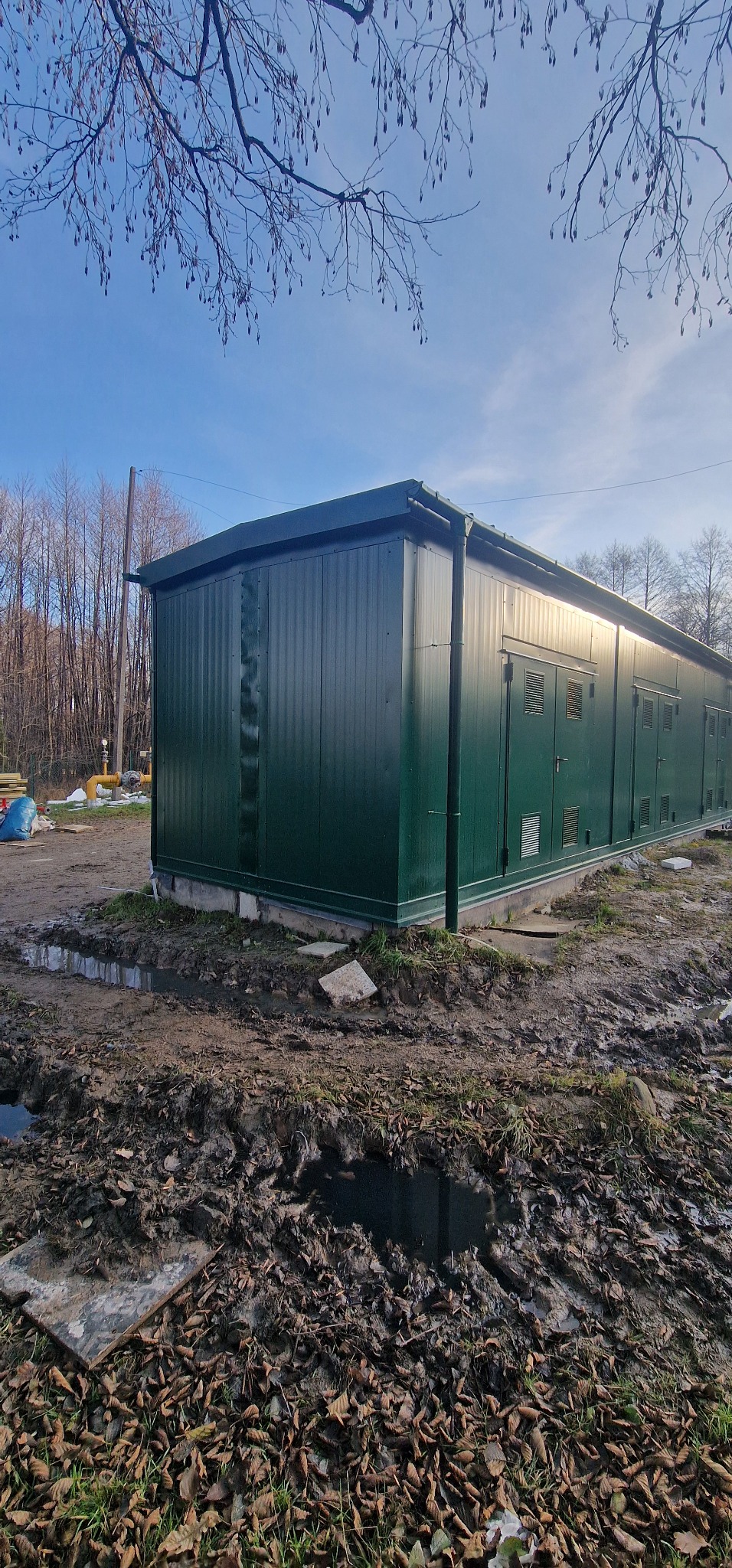 Zmontowany, długi pawilon z płyt warstwowych w kolorze ciemnej zieleni, z kilkoma drzwiami i kratkami wentylacyjnymi, otoczony błotnistym terenem i drzewami bez liści