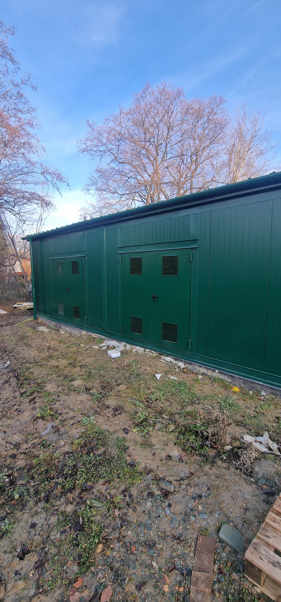 Gotowy, zielony pawilon z płyt warstwowych z dwoma parami drzwi z kratkami wentylacyjnymi, na nierównym, błotnistym terenie.