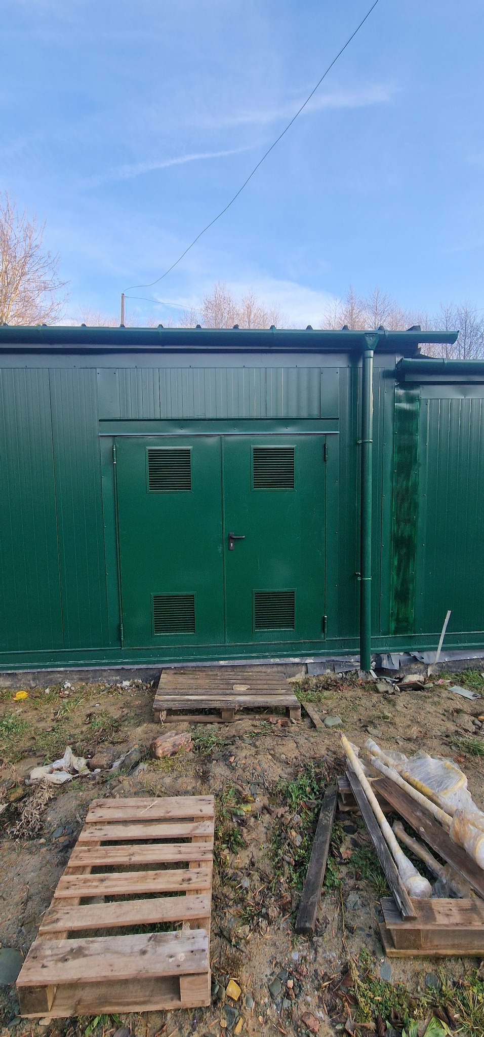 Zielony kontener z płyt warstwowych z podwójnymi drzwiami z kratkami wentylacyjnymi, stojący na ziemi z paletami i rurami w pobliżu.