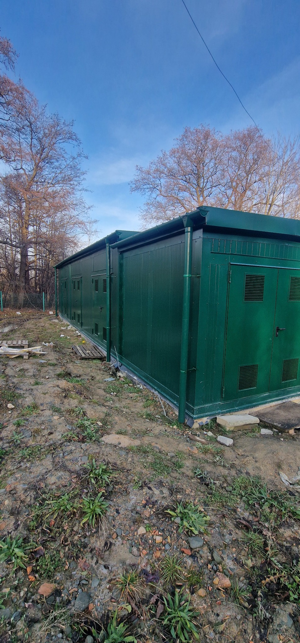 Zakończona budowa zielonej, modułowej konstrukcji z płyty warstwowej na fundamencie, widoczna z zewnątrz, z elementami wykończeniowymi, takimi jak rynny i drzwi z kratkami wentylacyjnymi, na tle...