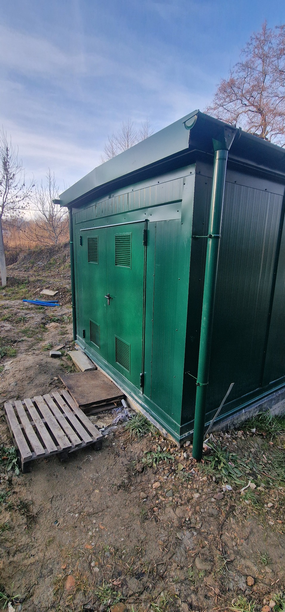 Zewnętrzna, zielona obudowa z płyt warstwowych z rynną, umieszczona na betonowej podstawie, z drewnianą paletą obok.