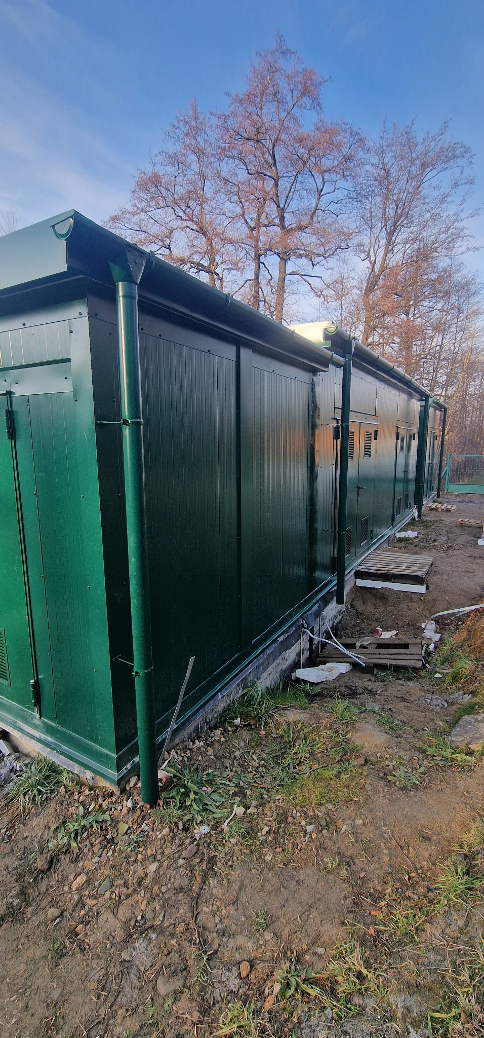 Długi, zielony pawilon z płyt warstwowych, z rynnami i drzwiami, ustawiony na fundamencie betonowym, w otoczeniu ziemi i drzew bez liści.