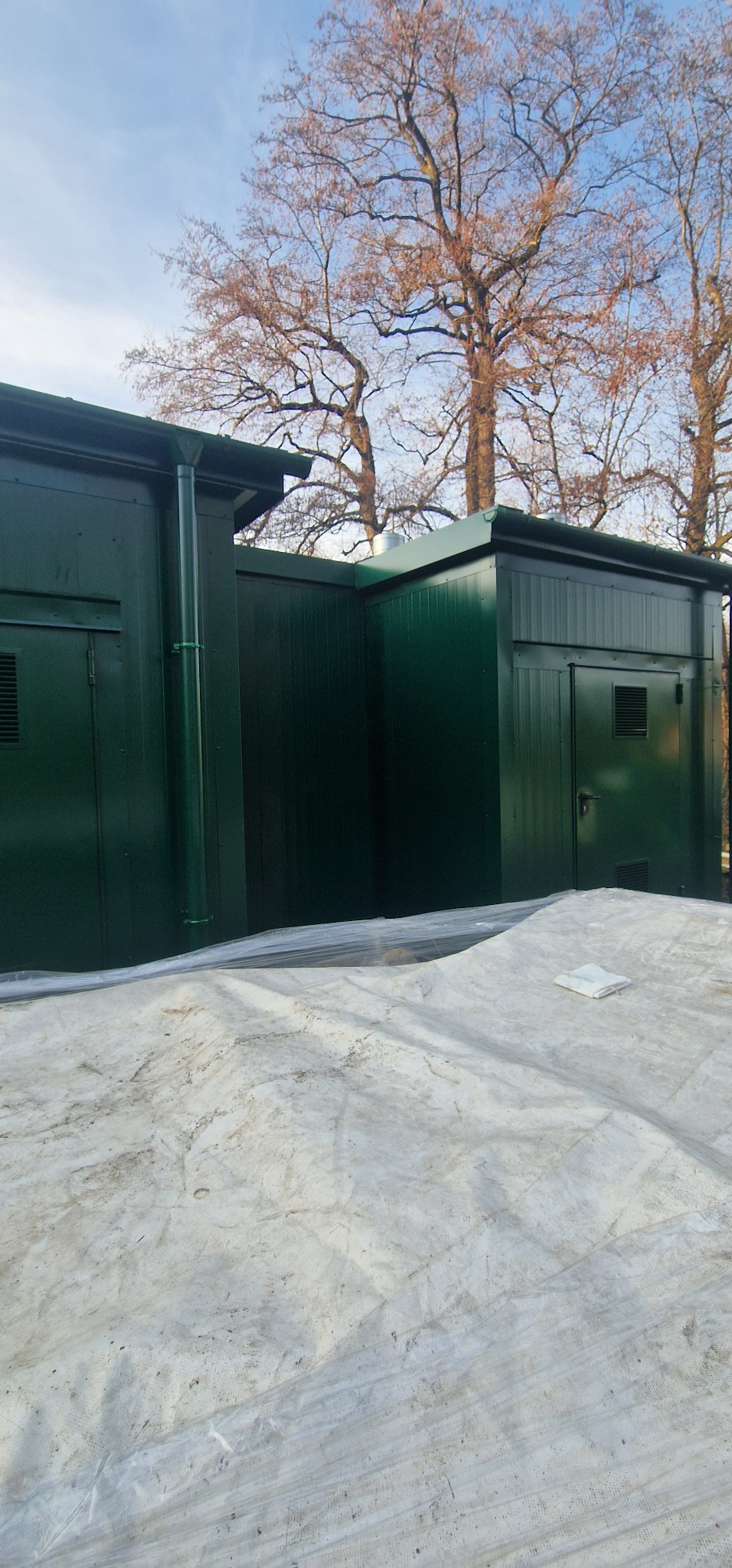 Zielony budynek z płyt warstwowych z widocznym orynnowaniem, zabezpieczony plandeką, na tle drzew bez liści i błękitnego nieba.