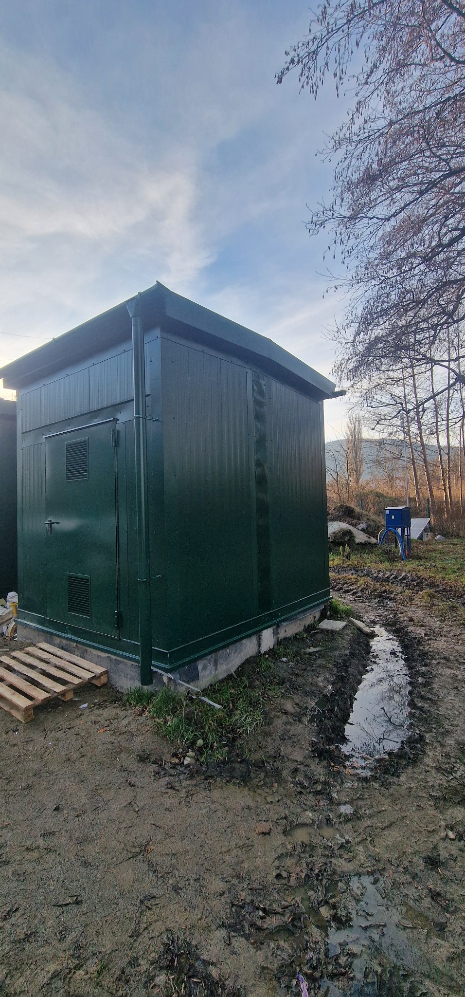 Gotowa, zielona budka z płyt warstwowych na betonowej podstawie, obok leżąca paleta, w tle drzewa i niebo.