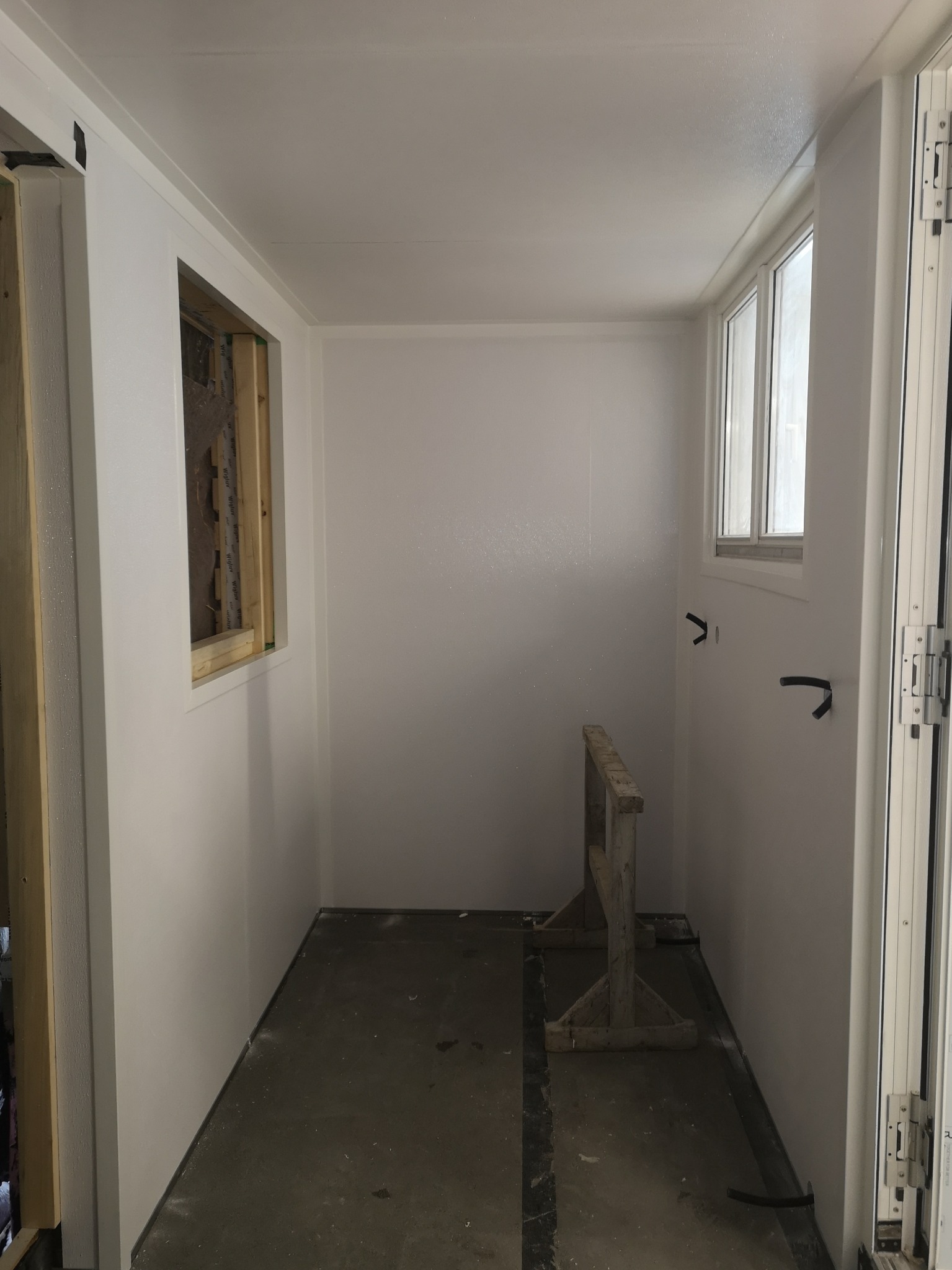 Wnętrze pomieszczenia z oknem i przygotowanym otworem okiennym, ściany wykończone na biało, widoczne ślady po pracach budowlanych na podłodze i drewniany kozioł.