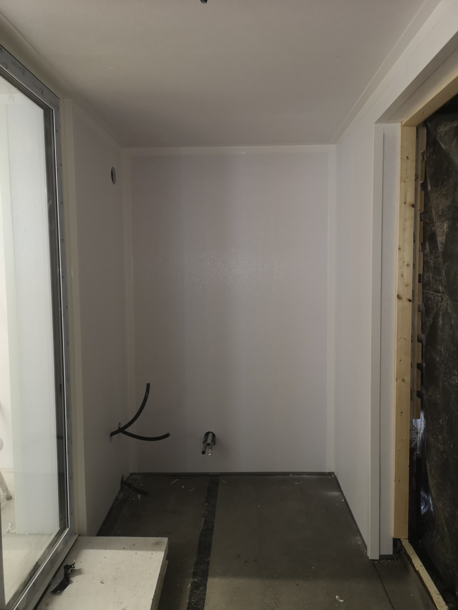 Wnętrze pomieszczenia w trakcie wykańczania w Rybniku: ściany wyłożone płytami, widoczne rury instalacji oraz fragment ramy okiennej i surowa ościeżnica drzwiowa.