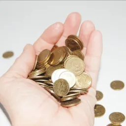 Otwarta dłoń trzymająca garść złotych monet, niektóre monety leżą na białym tle obok dłoni.