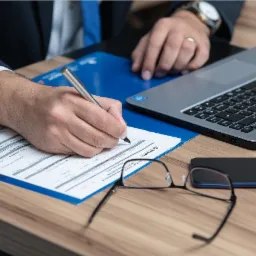 Osoba w garniturze wypełnia formularz długopisem na niebieskiej podkładce, obok leżą okulary i smartfon, w tle widoczny laptop.