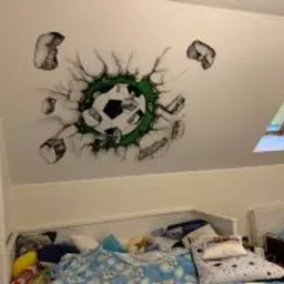 Malowidło na skosie sufitu w pokoju dziecięcym przedstawiające piłkę nożną przebijającą się przez ścianę, wykonane w czerni, bieli i zieleni, w tle okno dachowe i zasłona.