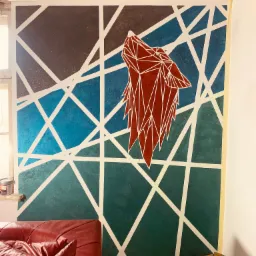 Ściana pomalowana w geometryczne wzory z motywem wilka w stylu origami, gradient kolorów od granatu do zieleni, białe linie przecinają tło, widoczny fragment czerwonej sofy i drewniany stołek.