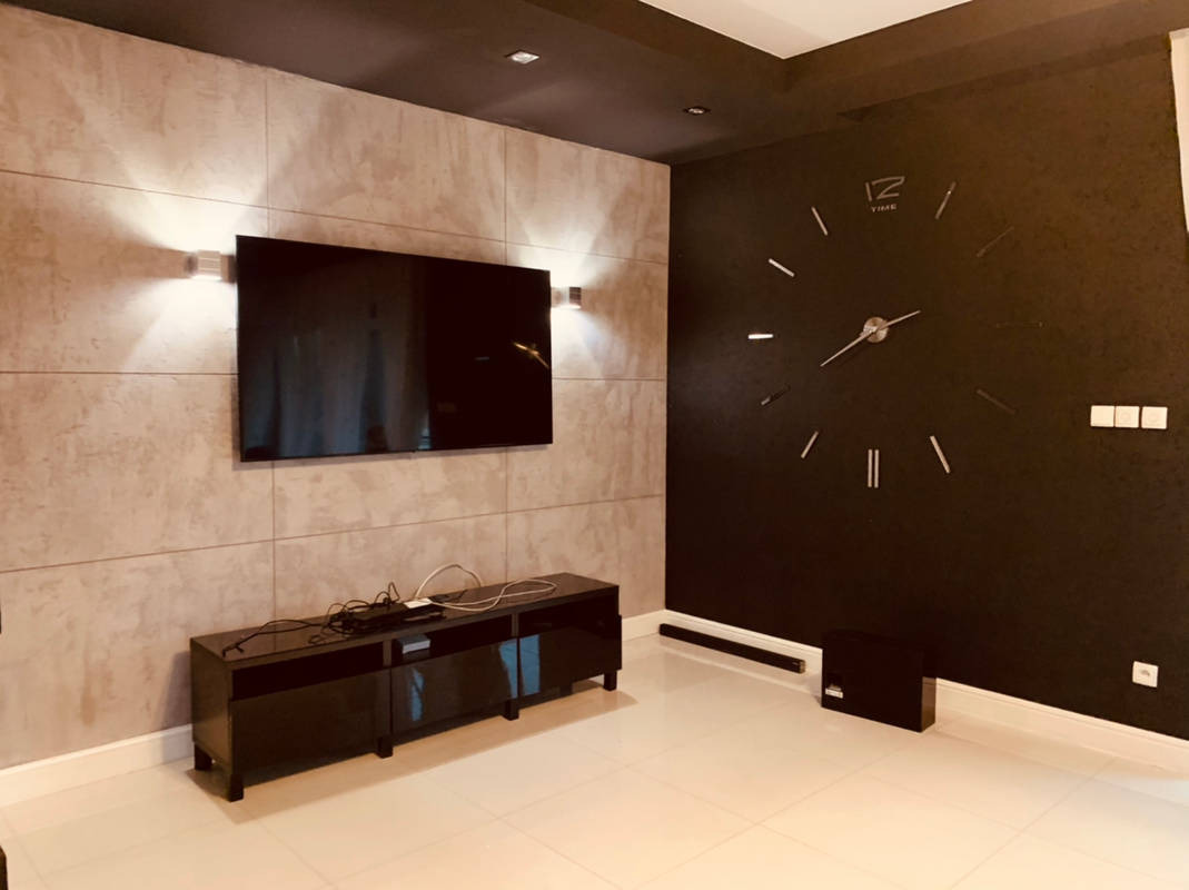 Nowoczesny salon z szarą ścianą o teksturze betonu, na której wisi telewizor podświetlony kinkietami, oraz czarną ścianą z dużym zegarem bez tarczy.