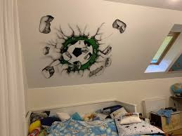 Malowidło na skosie sufitu w pokoju dziecięcym przedstawiające piłkę nożną przebijającą się przez ścianę, wykonane w czerni, bieli i zieleni, w tle okno dachowe i zasłona.