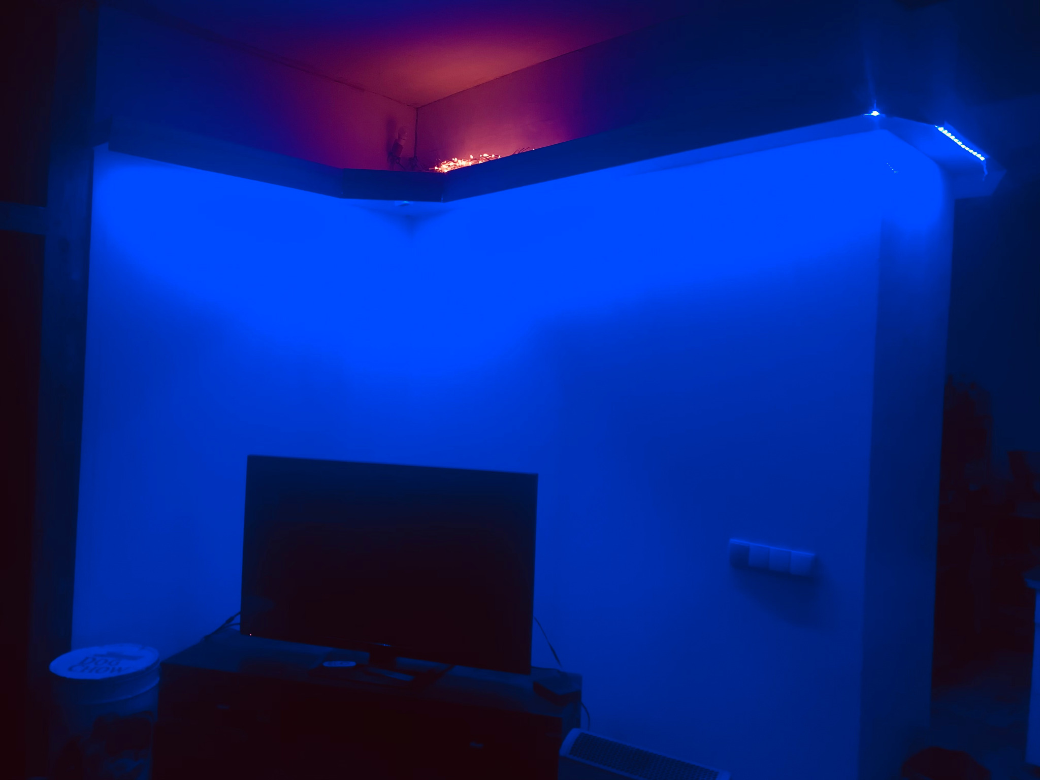 Pokój z pomalowanymi ścianami oświetlonymi niebieskim światłem LED, widoczny telewizor i elementy dekoracyjne.