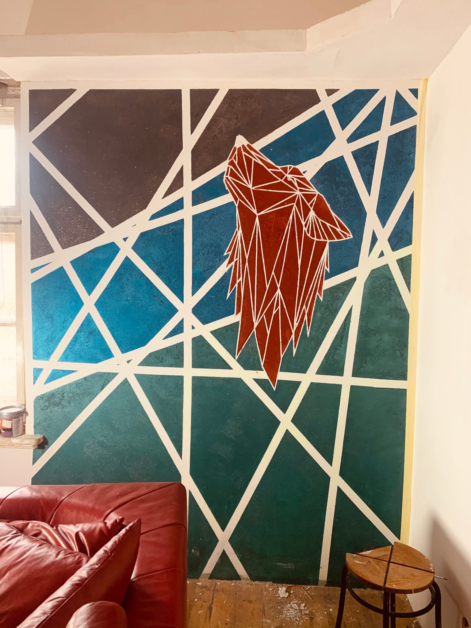Ściana pomalowana w geometryczne wzory z motywem wilka w stylu origami, gradient kolorów od granatu do zieleni, białe linie przecinają tło, widoczny fragment czerwonej sofy i drewniany stołek.