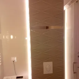 Beżowe płytki ułożone w romby z podświetleniem LED w wyremontowanej łazience, widoczny przycisk spłuczki i fragment pralki.