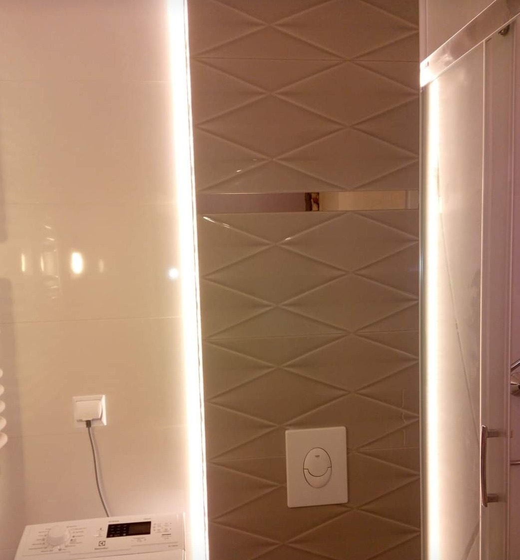 Beżowe płytki ułożone w romby z podświetleniem LED w wyremontowanej łazience, widoczny przycisk spłuczki i fragment pralki.