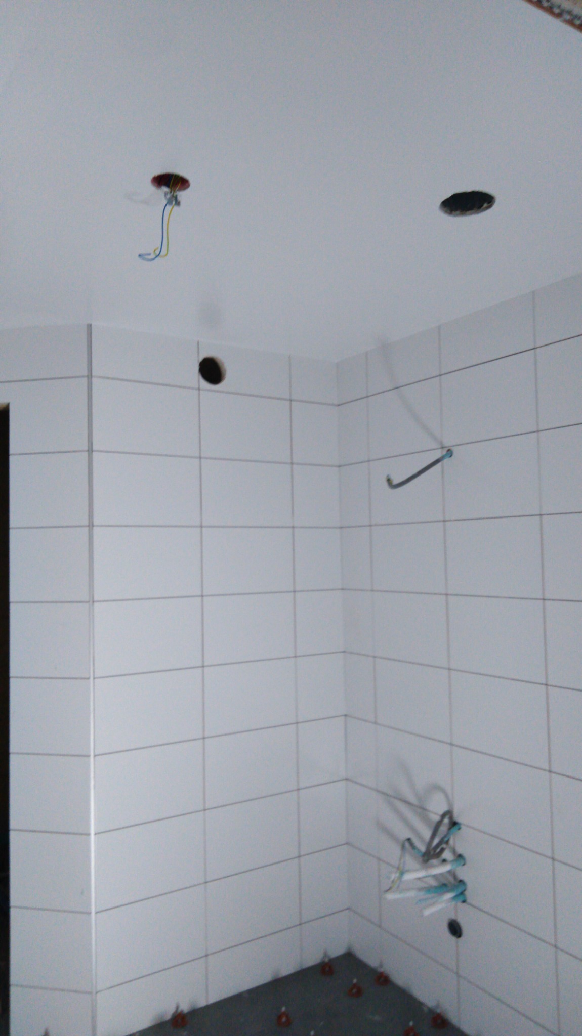 Wnętrze łazienki w trakcie prac wykończeniowych, widoczne białe płytki na ścianach, otwory na oświetlenie w suficie i ścianie, wystające przewody elektryczne oraz elementy poziomowania płytek...