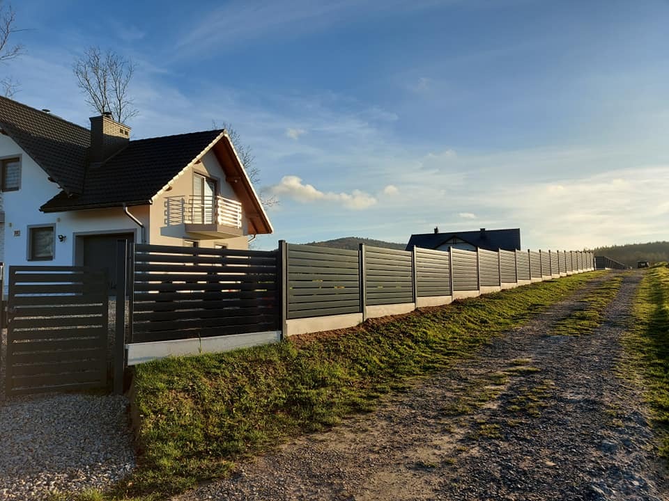 Nowoczesne, poziome ogrodzenie panelowe w kolorze antracytowym otacza posesję z białym domem, widoczne z polnej drogi w słoneczny dzień.