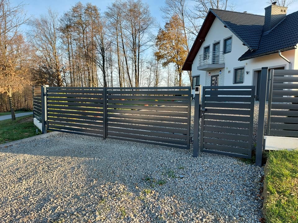 Nowoczesne, poziome przęsła ogrodzeniowe i brama w kolorze grafitowym, posesja z domem jednorodzinnym w tle, żwirowe podłoże.