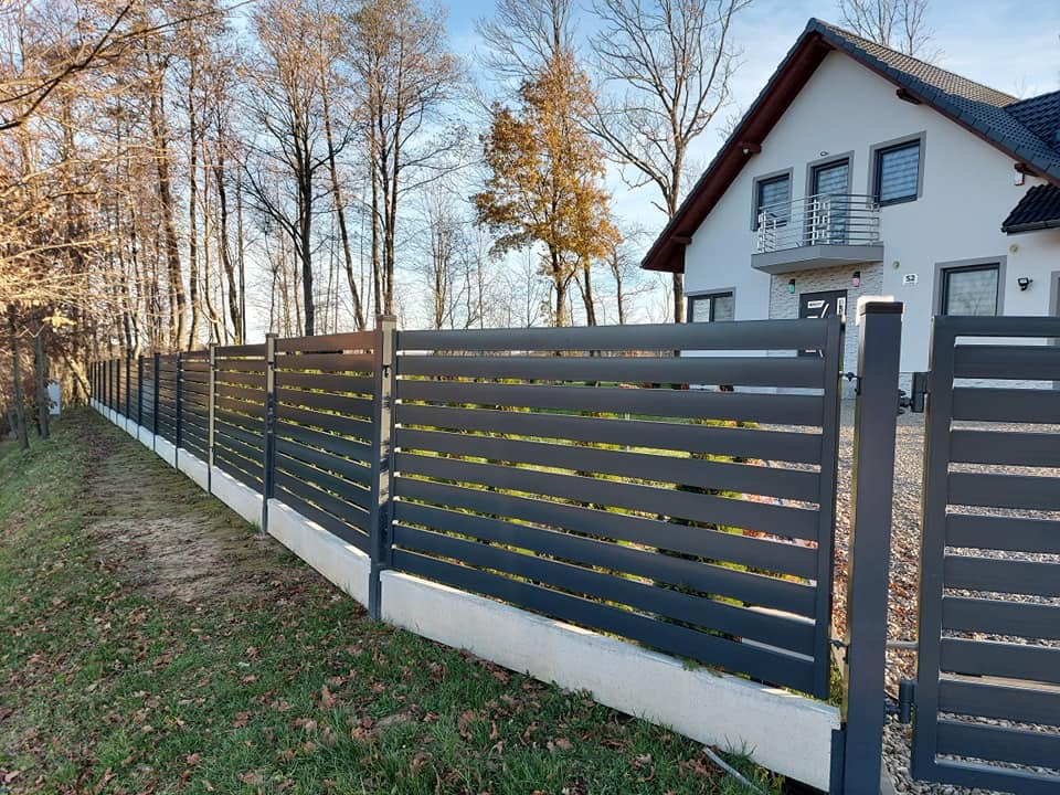 Nowoczesne, poziome ogrodzenie panelowe w kolorze grafitowym, na betonowej podmurówce, z widocznym domem w tle i drzewami bez liści.