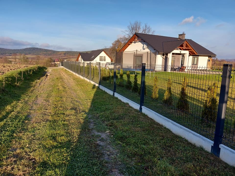 Długa, czarna, metalowa siatka ogrodzeniowa z betonową podmurówką, biegnąca wzdłuż polnej drogi, z młodymi tujami zasadzonymi po wewnętrznej stronie; w tle dwa nowe domy jednorodzinne z ciemnymi...