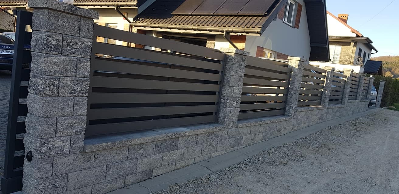 Nowoczesne ogrodzenie posesji z poziomymi, grafitowymi panelami, wsparte na słupkach z szarej kostki łupanej i podmurówce z tego samego materiału, na tle domu jednorodzinnego.