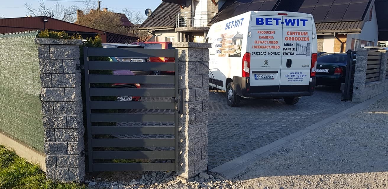 Nowoczesna, grafitowa brama z poziomymi panelami, osadzona na słupkach z szarej kostki łupanej; w tle zaparkowany biały samochód dostawczy z reklamą firmy ogrodzeniowej.