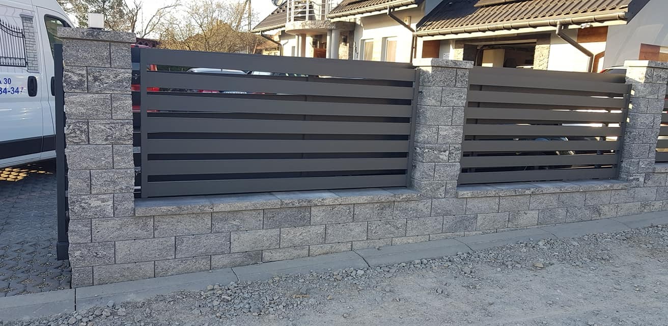 Nowoczesne ogrodzenie panelowe w kolorze grafitowym, z filarami i podmurówką z szarej kostki łupanej, widok z poziomu gruntu, na tle domu jednorodzinnego.