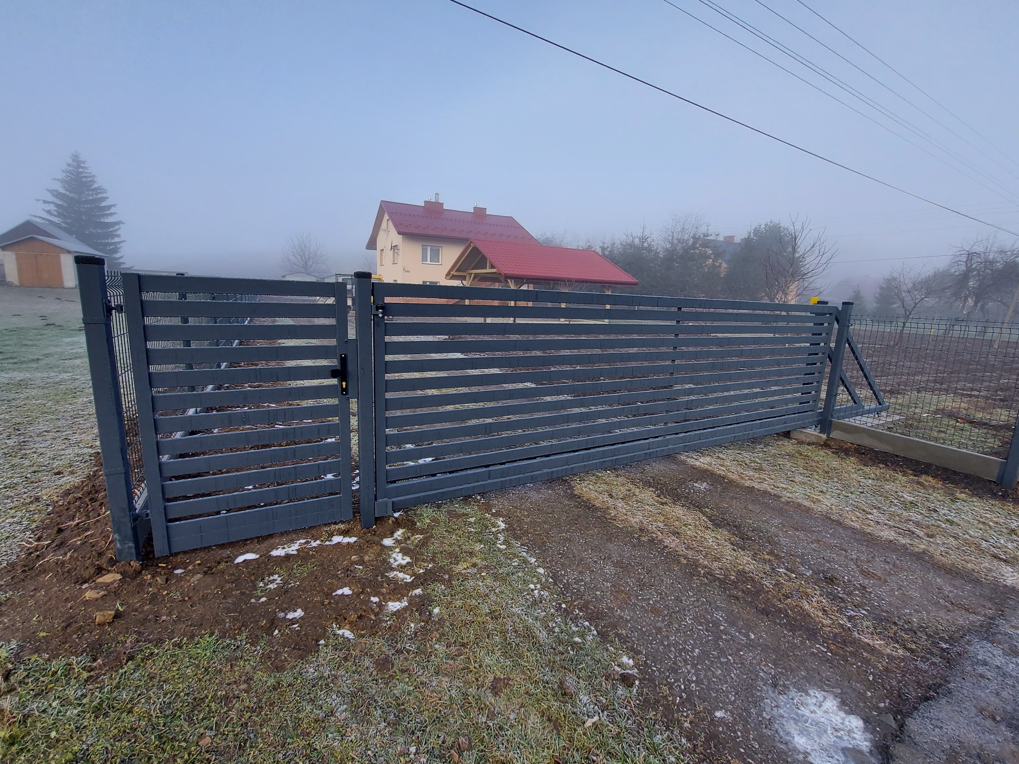 Nowoczesna, przesuwna brama wjazdowa z poziomych, szarych profili, częściowo otwarta, na tle zarośniętego terenu i domu z czerwonym dachem w porannej mgle.