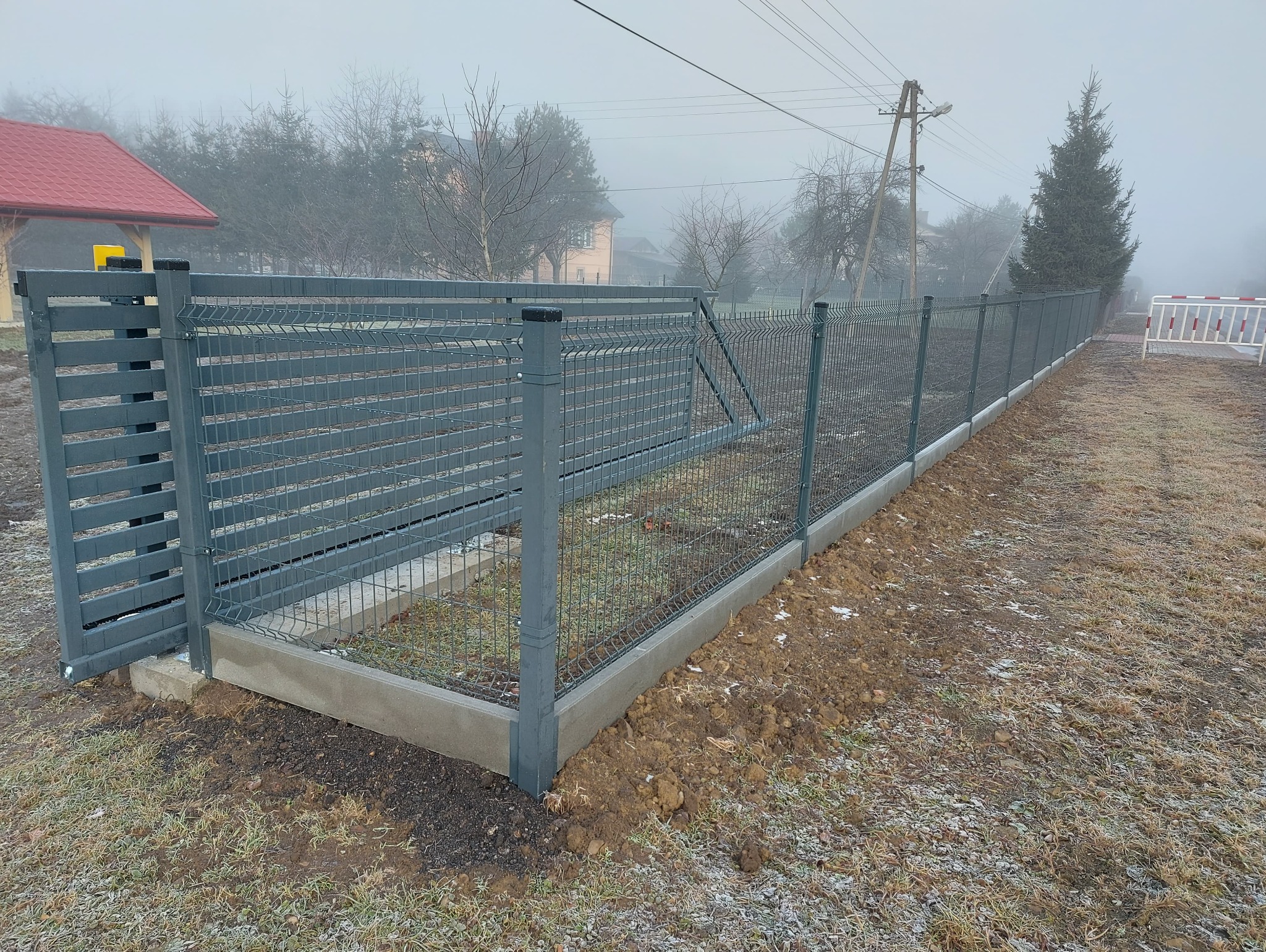 Nowo zainstalowane, szare ogrodzenie panelowe z betonową podmurówką w pochmurny dzień, widoczny fragment posesji i budynki w tle.