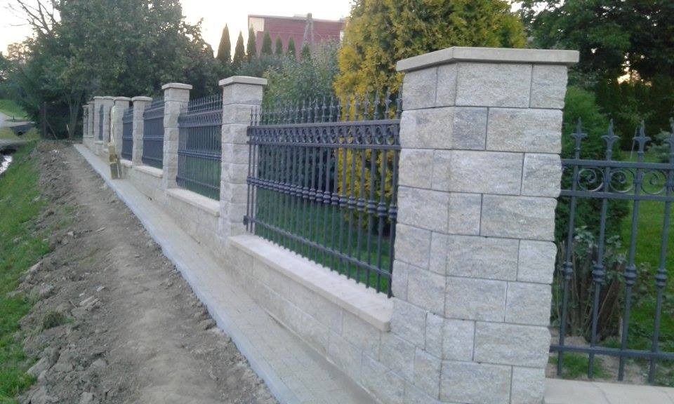 Nowoczesne ogrodzenie z szarego kamienia łupanego z metalowymi przęsłami, widok z boku, za ogrodzeniem zieleń drzew i krzewów, w tle budynek z czerwonym dachem.