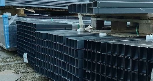 Stosy profili kwadratowych z metalu w kolorze grafitowym, ułożone na paletach i zabezpieczone folią stretch, przygotowane do transportu lub magazynowania.