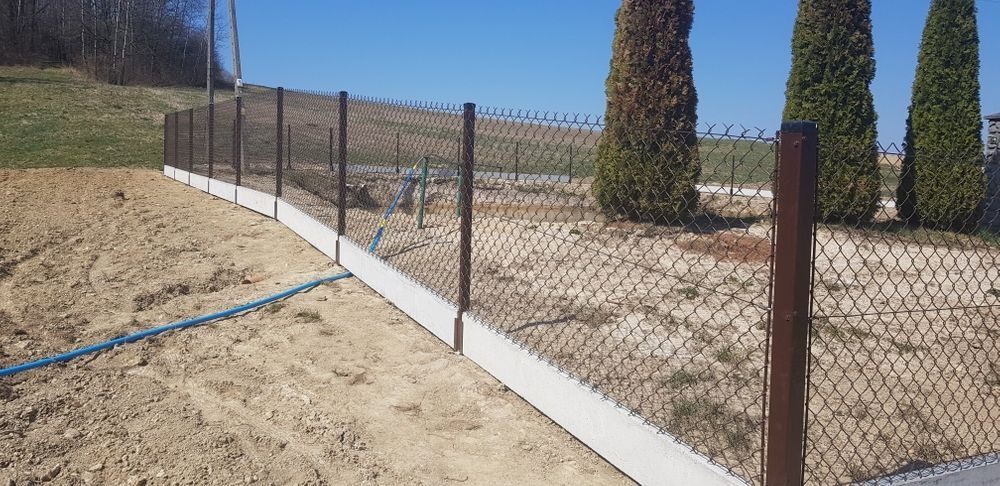 Ogrodzenie z siatki z brązowymi słupkami i betonową podmurówką na nierównym terenie, widoczny fragment niebieskiego węża ogrodowego.