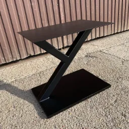 Czarna, metalowa podstawa stolika z geometrycznym spawem, widoczna na tle brązowej blachy falistej i betonowej nawierzchni.