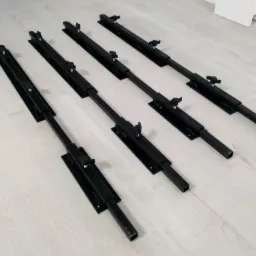 Cztery czarne, metalowe rygle do bram przemysłowych z regulowanymi pokrętłami, ułożone równolegle na jasnej podłodze z paneli.