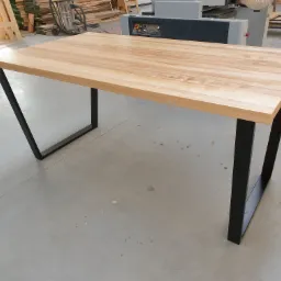 Gotowy stół z blatem z jasnego drewna i czarnymi, metalowymi nogami w kształcie trapezu, widoczny w warsztacie na betonowej podłodze, z maszynami do obróbki drewna w tle.