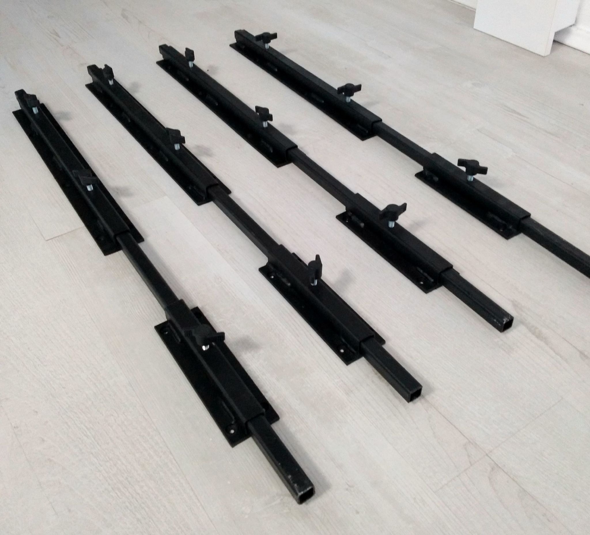 Cztery czarne, metalowe rygle do bram przemysłowych z regulowanymi pokrętłami, ułożone równolegle na jasnej podłodze z paneli.