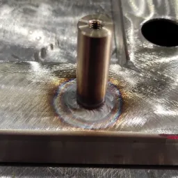 Metalowa podstawa z przyspawanym, mosiężnym elementem cylindrycznym z gwintem wewnętrznym, widoczne ślady spawania i obr&oacute;bki.