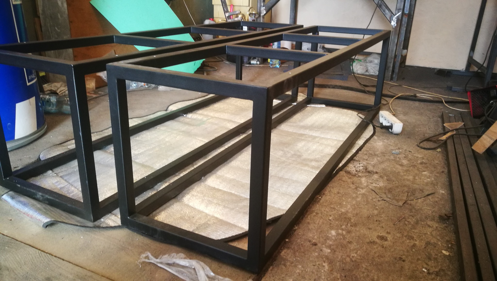 Dwie czarne, spawane ramy z profili stalowych, prawdopodobnie przygotowane do dalszej obróbki lub montażu, leżące na podłodze warsztatu, widoczny przedłużacz i resztki materiałów.